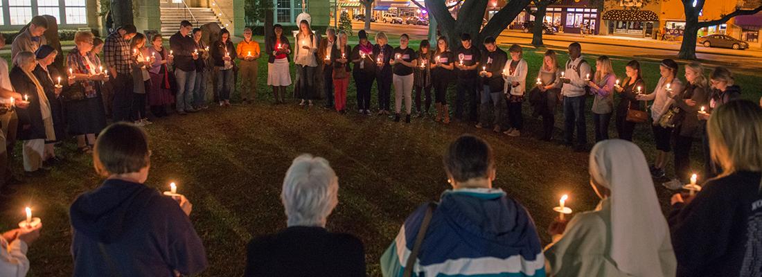 A circle of people holding candles gather for a nighttime prayer vigil on the lawn of the TCU campus.